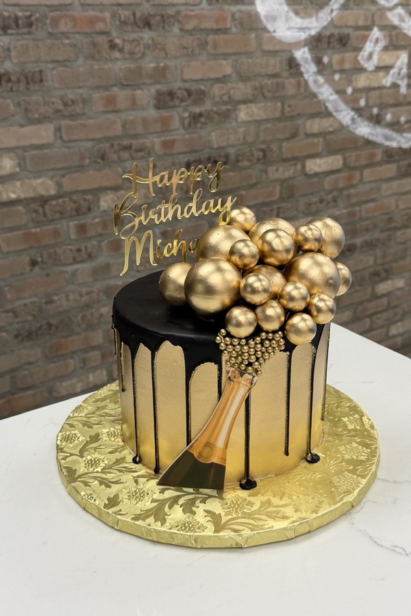Black and gold birthday cake with chocolate drip, gold bubbles, champagne bottle and personalized topper.