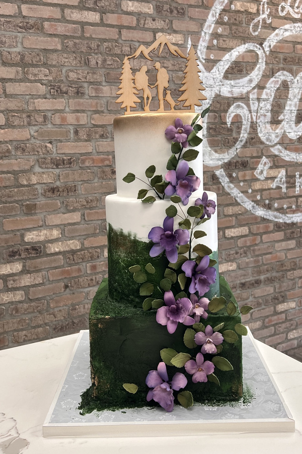 Nature-inspired wedding cake with purple flowers, cascading leaves, mountain scenery and hiking couple topper.