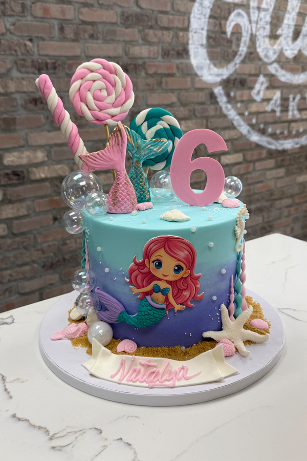Mermaid sixth birthday cake with mermaid tails, lollipops, bubbles, starfish, seashells and number 6.
