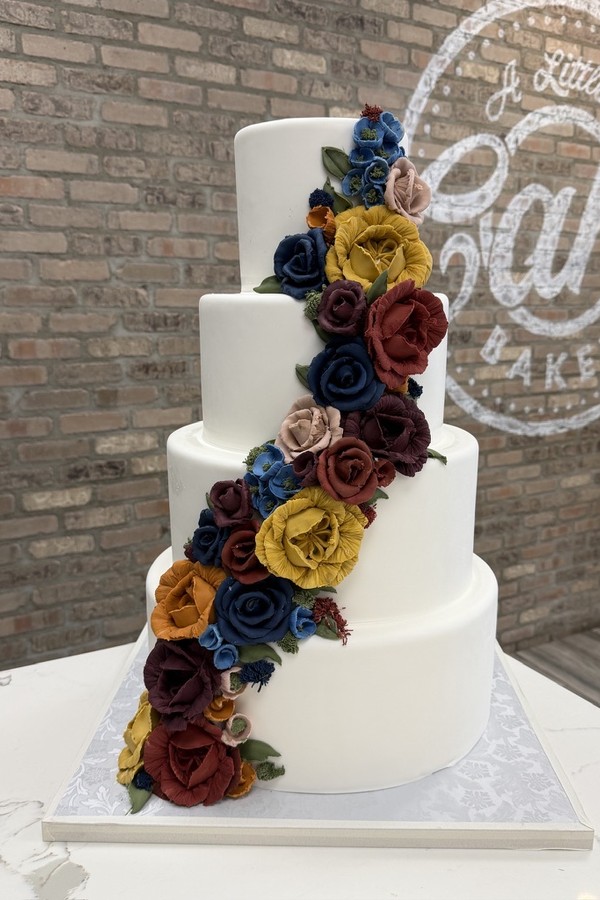 Four-tier white wedding cake with cascading burgundy, navy, yellow, orange, pink and red sugar flowers.