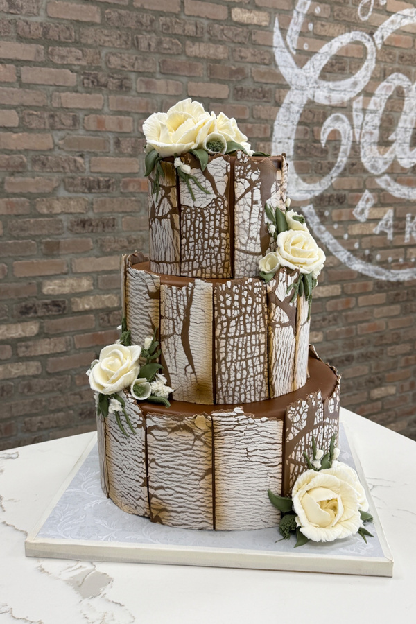 Three-tier chocolate wedding cake with textured bark panels, ivory roses, flowers and greenery.