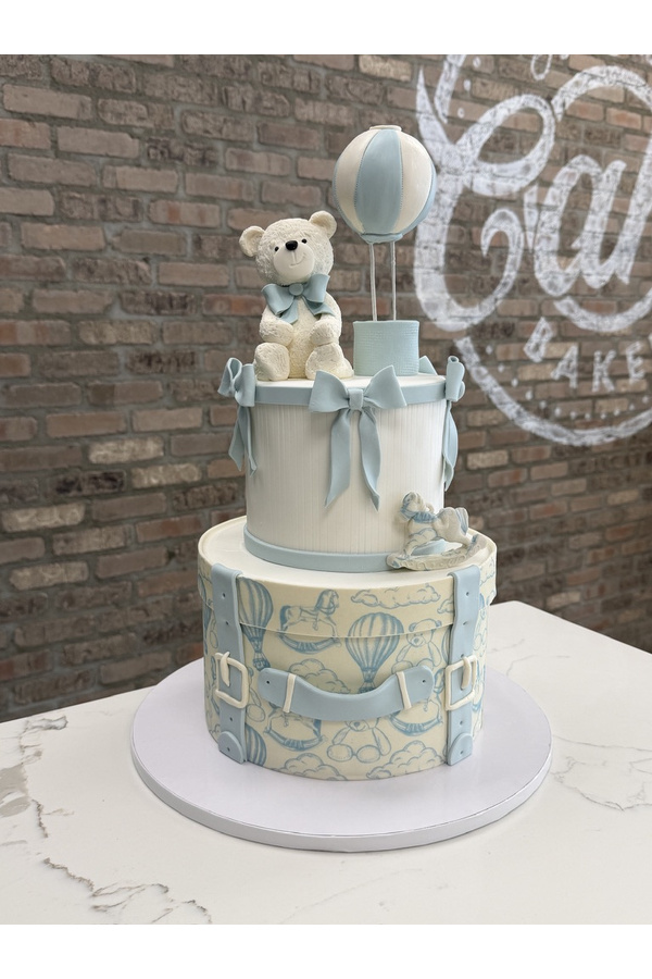 Two-tier baby shower cake with teddy bear, hot air balloon, rocking horse and vintage suitcase design.