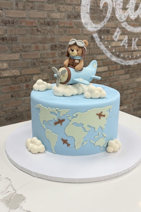 Blue travel cake with teddy bear pilot, airplane, clouds, world map and flight path details.
