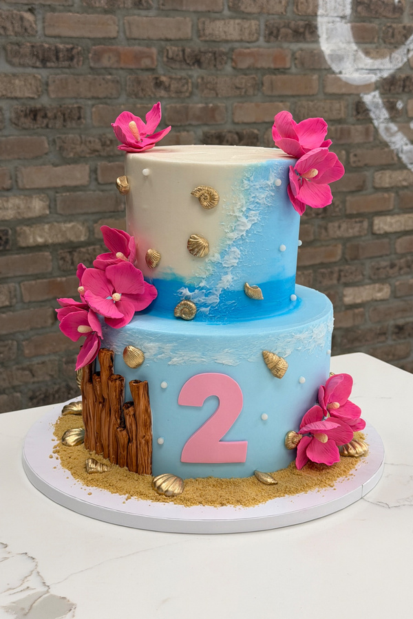 Two-tier beach birthday cake with ocean waves, seashells, tropical flowers, sand and number 2.