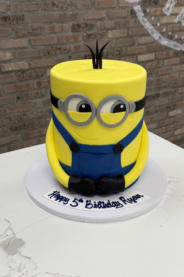 Sculpted yellow Minion birthday cake with silver goggles, blue overalls and black boots.